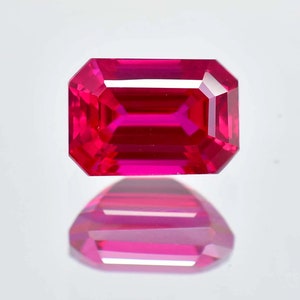 May include: A vibrant, deep red emerald-cut gemstone with a high polish. The gemstone is reflecting light, creating a bright, shiny effect.