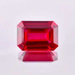 May include: A deep red emerald cut gemstone with a high polish. The gemstone is reflecting light and has a slight shadow underneath.