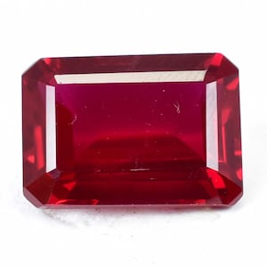 May include: A deep red gemstone with a rectangular emerald cut. The gemstone is faceted and has a high polish.