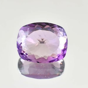 May include: A faceted, oval-shaped amethyst gemstone. The gem features a gradient of colors, transitioning from a deep purple at the edges to a lighter lavender hue in the center. The stone is highly reflective, with a polished surface.