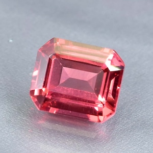 May include: A vibrant red gemstone with a rectangular emerald cut. The gemstone has a slight pink hue and is cut with precision.