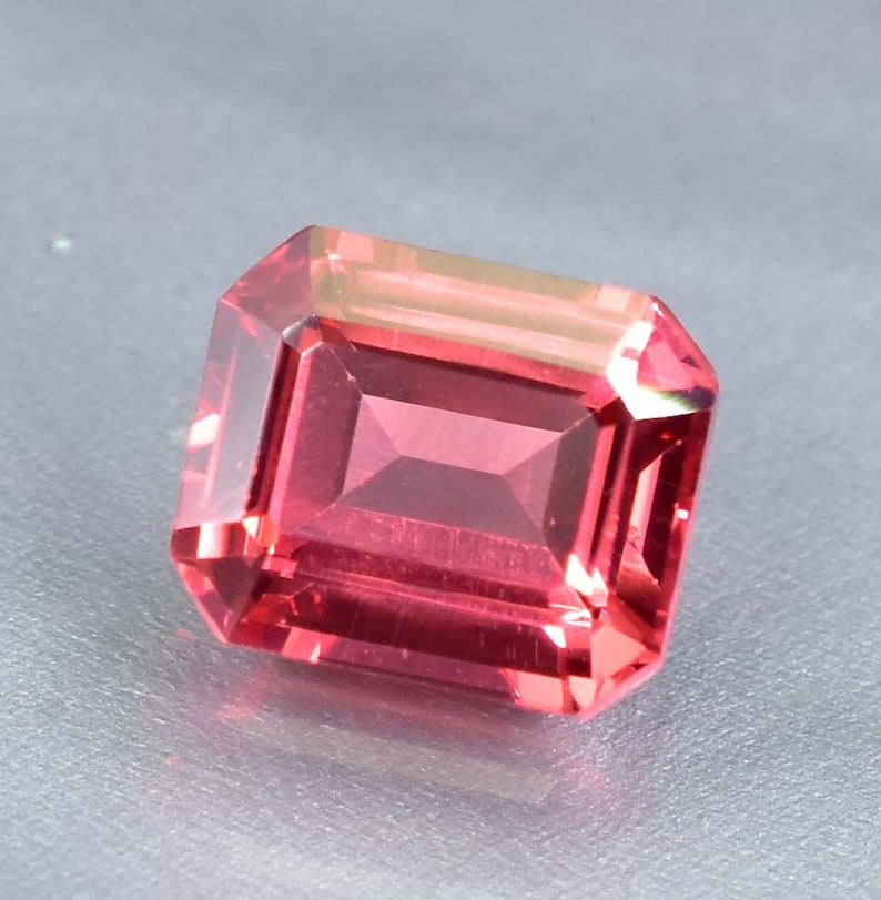May include: A vibrant red gemstone with a rectangular emerald cut. The gemstone has a slight pink hue and is cut with precision.