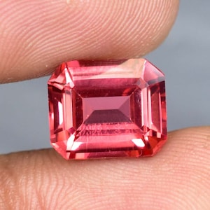 May include: A vibrant pink gemstone with a rectangular emerald cut. The gemstone is faceted and has a high polish.