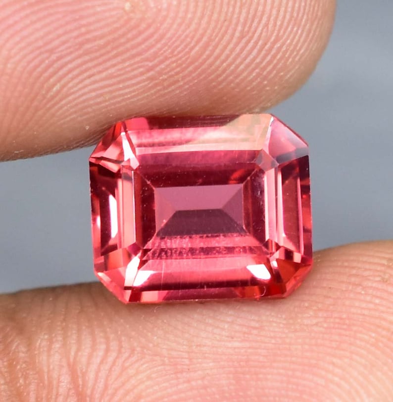 May include: A vibrant pink gemstone with a rectangular emerald cut. The gemstone is faceted and has a high polish.