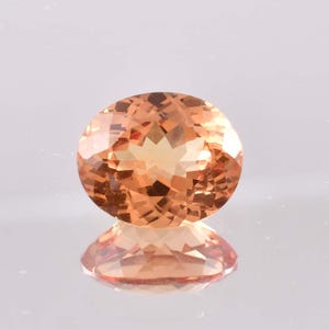 May include: An oval-cut gemstone with a peach-colored hue. The gem is faceted, reflecting light and creating a sparkling effect. The stone is set against a reflective surface, showing its mirrored image.
