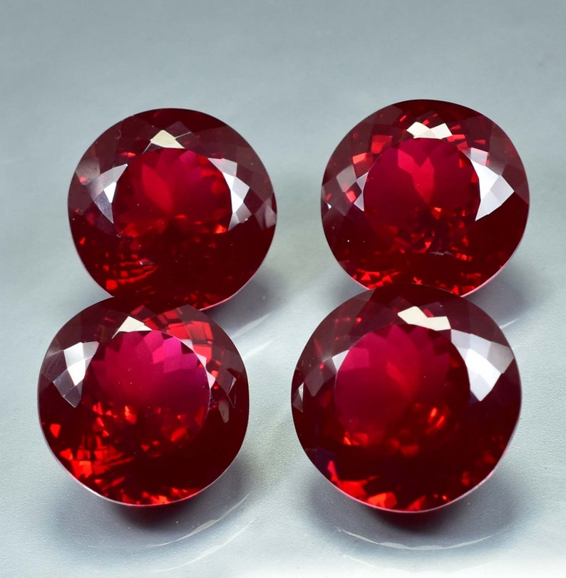 Pode incluir: Quatro pedras preciosas redondas de cor vermelho escuro com corte facetado. As pedras preciosas est&atilde;o dispostas em forma de quadrado sobre uma superf&iacute;cie cinza.