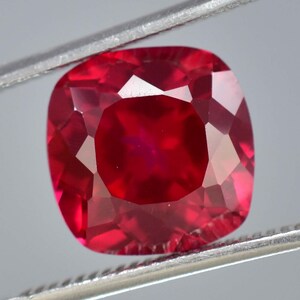 May include: A deep red gemstone with a cushion cut. The gemstone is held in a pair of tweezers.