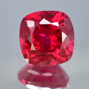 May include: A deep red gemstone with a cushion cut. The gemstone is faceted and has a high polish.