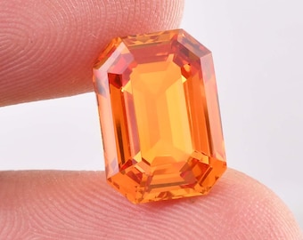 May include: A faceted, rectangular-cut gemstone with a vibrant orange hue. The gem's facets reflect light, creating a brilliant display. The gemstone is held between two fingers, showcasing its size and shape. This gemstone is suitable for jewelry making.