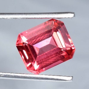 May include: A vibrant pink gemstone with a rectangular emerald cut. The gemstone is held in place by a pair of tweezers.