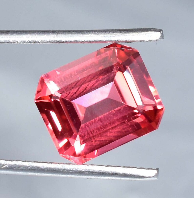 May include: A vibrant pink gemstone with a rectangular emerald cut. The gemstone is held in place by a pair of tweezers.