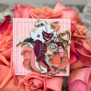 Puede incluir: Un pin de esmalte dorado con dos personajes de dibujos animados abrazándose. Los personajes son en tonos blanco, burdeos, rosa y negro, con un elemento en forma de corazón. El pin está sobre un fondo rosa a rayas y rodeado de rosas rosas.