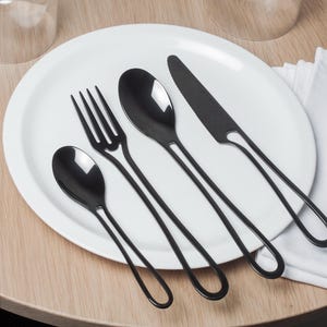 K&ouml;nnte beinhalten: Ein Set schwarzes Besteck auf einem wei&szlig;en Teller. Das Set enth&auml;lt eine Gabel, ein Messer und zwei L&ouml;ffel.