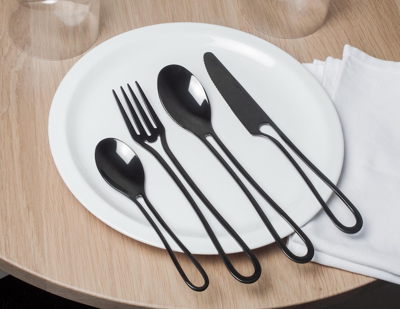 K&ouml;nnte beinhalten: Ein Set schwarzes Besteck auf einem wei&szlig;en Teller. Das Set enth&auml;lt eine Gabel, ein Messer und zwei L&ouml;ffel.