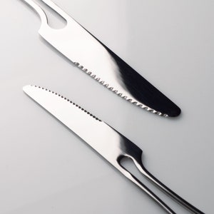 K&ouml;nnte beinhalten: Zwei silberne gezackte Messer mit unterschiedlichen Griffen. Das obere Messer hat einen einfachen Griff mit einem Loch in der Mitte. Das untere Messer hat einen zweizinkigen Griff.