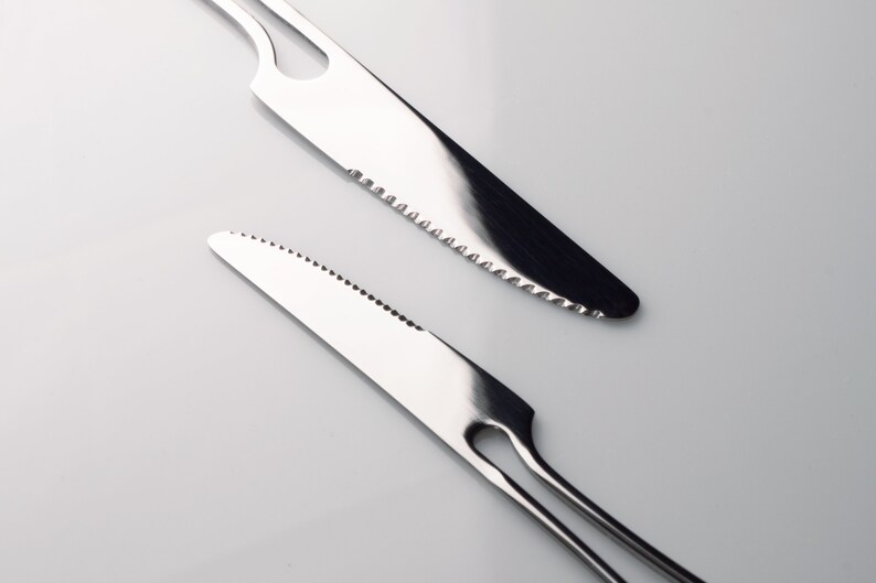 K&ouml;nnte beinhalten: Zwei silberne gezackte Messer mit unterschiedlichen Griffen. Das obere Messer hat einen einfachen Griff mit einem Loch in der Mitte. Das untere Messer hat einen zweizinkigen Griff.