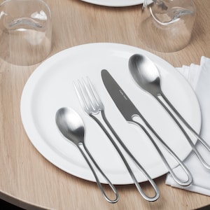K&ouml;nnte beinhalten: Ein wei&szlig;er Teller mit einer Gabel, einem Messer und zwei L&ouml;ffeln. Das Besteck hat ein einzigartiges, geschwungenes Design. Der Teller steht auf einem Holztisch mit einer wei&szlig;en Serviette.
