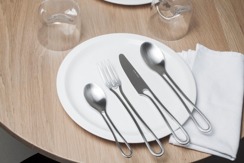 K&ouml;nnte beinhalten: Ein wei&szlig;er Teller mit einer Gabel, einem Messer und zwei L&ouml;ffeln. Das Besteck hat ein einzigartiges, geschwungenes Design. Der Teller steht auf einem Holztisch mit einer wei&szlig;en Serviette.