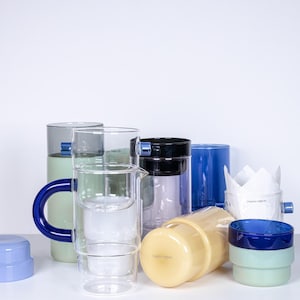 Op de afbeelding: Een verzameling glazen en keramische bekers en kannen in verschillende kleuren, waaronder blauw, groen, geel en wit. De bekers en kannen zijn gestapelt en gerangschikt op een wit oppervlak.