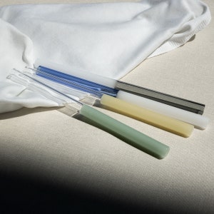Puede incluir: Cuatro pajitas para beber de vidrio de diferentes colores: verde, amarillo, blanco y azul. Las pajitas están sobre una superficie marrón claro.