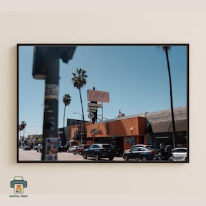 May include: A framed digital print depicting a street scene with a restaurant sign, palm trees, and parked cars against a clear blue sky. The restaurant sign reads "Canter's".