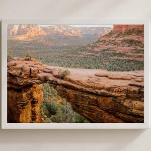 May include: A framed landscape photograph of a natural rock arch in a desert environment. The arch is a reddish-brown color, with a valley of green trees and distant mountains in the background. The photograph is in a white frame.