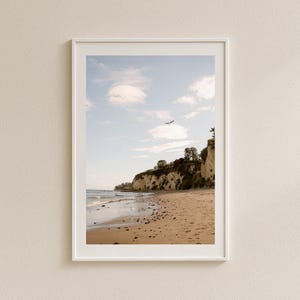 Può includere: Fotografia incorniciata di un paesaggio costiero. L'immagine presenta una spiaggia sabbiosa, una scogliera con vegetazione e un cielo blu con nuvole. Un uccello vola nel cielo. La foto è in una cornice bianca.