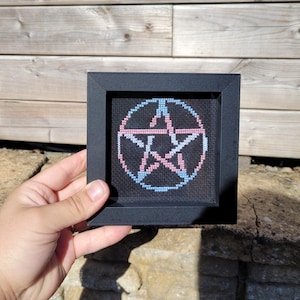May include: A framed cross-stitch of a pentagram with a pink and blue design. The pentagram is surrounded by a circle.