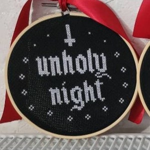 May include: A black cross-stitch hoop with the words "unholy night" in white. The design includes a cross symbol and small star-like accents. A red ribbon is attached for hanging. The hoop is set against a white wall.