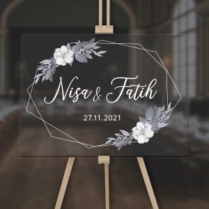 Könnte beinhalten: Ein schwarzes Acryl-Hochzeitschild mit einem weißen geometrischen Rahmen und floralem Design. Das Schild trägt die Aufschrift "Nisa & Fatih" und "27.11.2021".