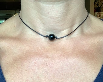 May include: A black choker necklace featuring a single, round, dark bead. The bead is centered on a thin, black cord. The necklace is worn against a person's skin, with a dark blue v-neck shirt visible in the frame.