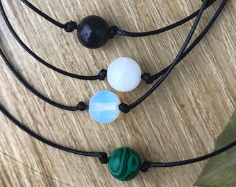 May include: Four necklaces with different colored beads. The necklaces are made of black string and have a single bead on each. The beads are purple, white, light blue, and green.