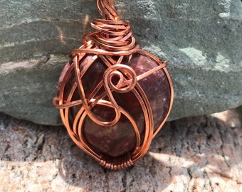 May include: A copper wire wrapped pendant featuring a red and brown jasper stone. The pendant is hanging from a black cord.