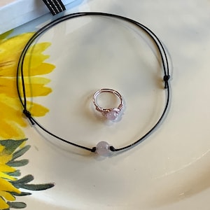 May include: A delicate pink stone ring and a simple black cord necklace with a pink stone bead. Both pieces of jewelry are on a white surface.