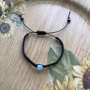 May include: A black braided bracelet with a light blue opal bead. The bracelet is tied with black cord and has black beads at the ends.