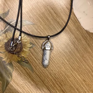 May include: A white and gray stone pendant with a silver setting hangs from a black leather cord necklace.