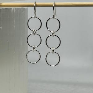 May include: A pair of silver earrings with a cascading three-circle design. Each earring features three connected, open circles suspended from a hook. The earrings are displayed against a neutral background, highlighting their minimalist style.