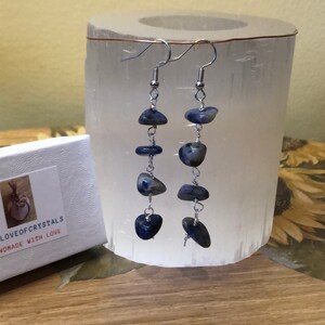 May include: Dangle earrings featuring dark blue, irregularly shaped stones strung on silver-colored wire. The earrings are displayed next to a white box with the text "LOVE OF CRYSTALS HANDMADE WITH LOVE". The background includes a white cylindrical object and a wooden surface.