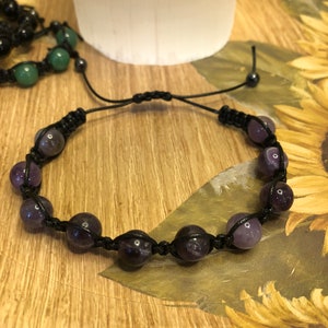 May include: A black cord bracelet with dark purple gemstone beads. The bracelet is tied with a knot and has a simple design.