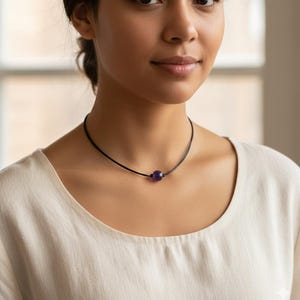 May include: A black choker necklace featuring a single, round, deep purple amethyst gemstone. The gemstone is centered on a thin, black cord. The necklace is worn around the neck.