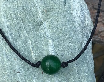 May include: A dark green stone bead on a black cord necklace. The necklace is displayed on a light gray rock.