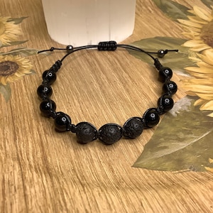 May include: Black beaded bracelet featuring polished onyx and porous lava stones. The bracelet is strung on a black cord with an adjustable sliding knot closure. The bracelet's beads are approximately 0.4 inches in diameter.