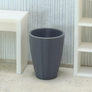 Puede incluir: Un cubo de basura gris y cilíndrico con una superficie texturizada. El cubo de basura está en una habitación con un escritorio, una silla y una estantería blancos. El fondo tiene un papel pintado a rayas azul claro y blanco.
