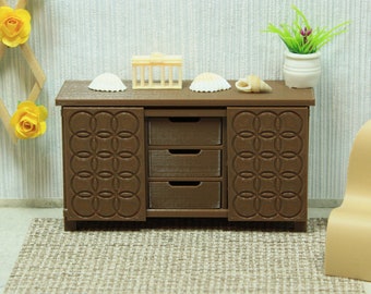 Puede incluir: Un armario de madera marrón con tres cajones y dos puertas. Las puertas tienen un patrón circular tallado en ellas. El armario está sobre una alfombra marrón. Hay conchas marinas y un pequeño objeto decorativo en la parte superior del armario.