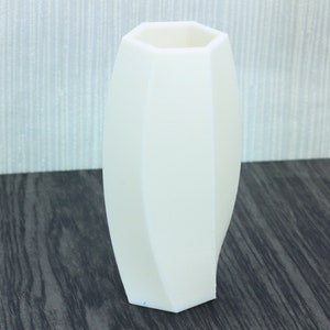 Puede incluir: Un jarrón de cerámica blanco con forma geométrica octogonal. El jarrón está sobre una superficie de madera oscura.