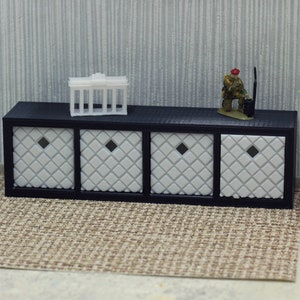 May include: A black wooden shelf with four white drawers featuring a diamond pattern. The shelf is placed on a brown rug. There is a small white model building and a miniature figurine on top of the shelf.