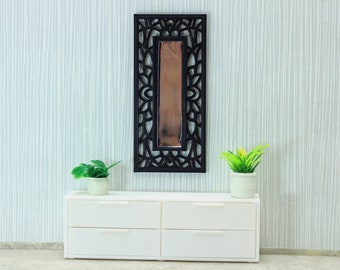 May include: A white dresser with four drawers sits in front of a white wall with a black decorative mirror with a geometric design above it. Two potted plants are on either side of the dresser.