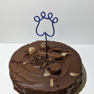 May include: A round chocolate cake with chocolate frosting, decorated with chocolate shavings, candies, and a blue paw print cake topper. The cake is on a white surface.