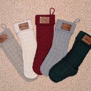 Puede incluir: Cinco calcetines navideños de punto trenzado en varios colores: gris, crema, burdeos, gris claro y verde oscuro. Cada calcetín tiene una etiqueta de cuero con un nombre: Beth, Brody, Darcy, Duncan y Zachary. Fondo neutro.