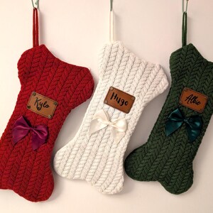May include: Three knitted dog bone-shaped Christmas stockings in red, white, and green. Each stocking has a leather name tag and a satin bow. The red stocking has a burgundy bow, the white has a cream bow, and the green has a dark green bow.